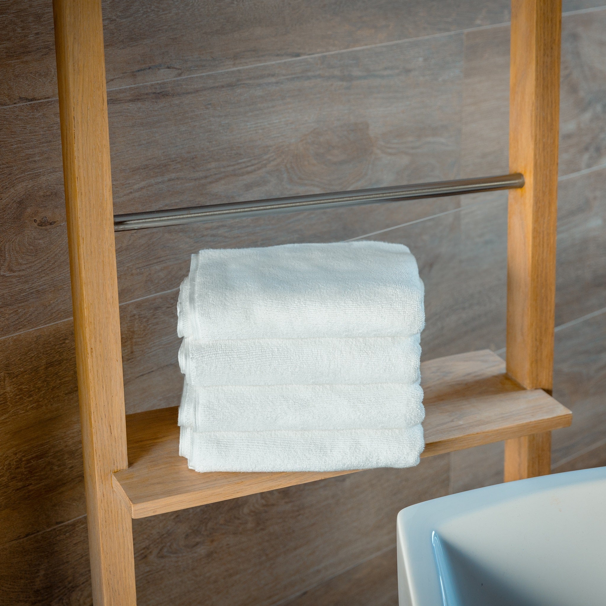 Stack of white bathmats on a wooden towel rack against a tiled wall.