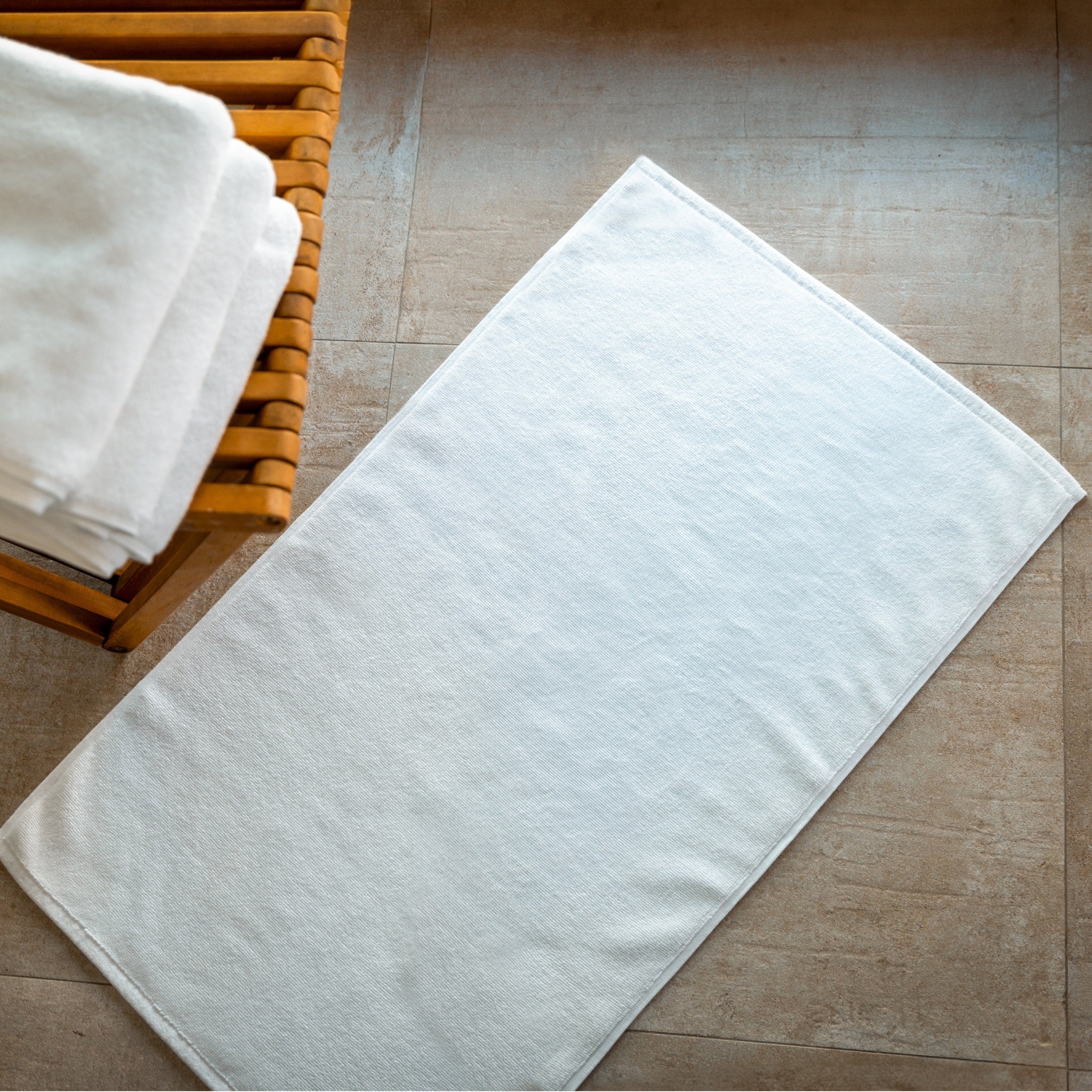 White bath mat on a tiled bathroom floor with a wooden basket containing additional white bath mats.