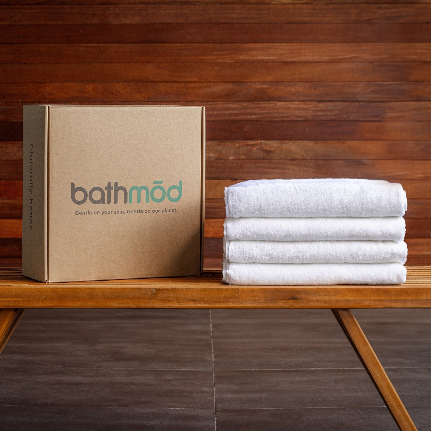 Stack of white towels next to a brown 'bathmöd' box on a wooden surface with a wooden wall background.