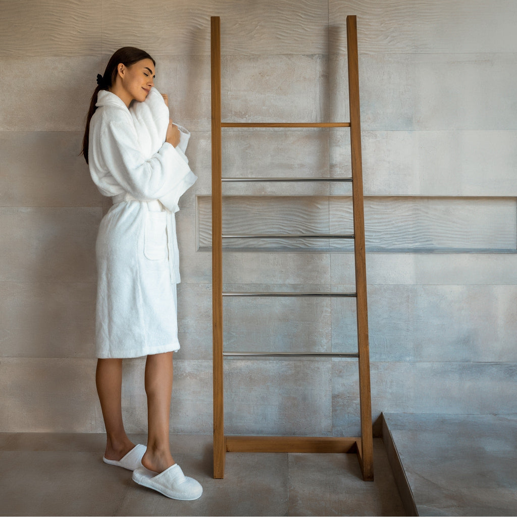 Woman in a white robe standing next to a wooden towel ladder in a bathroom.