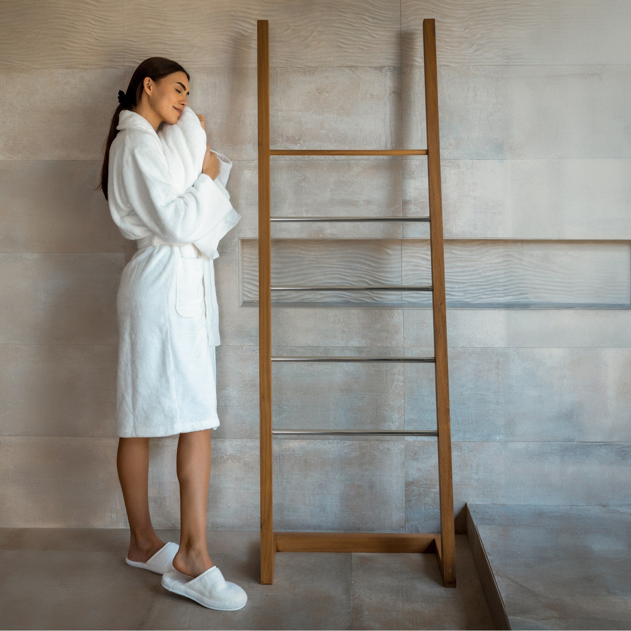 Woman in a white robe standing next to a wooden towel ladder in a bathroom.