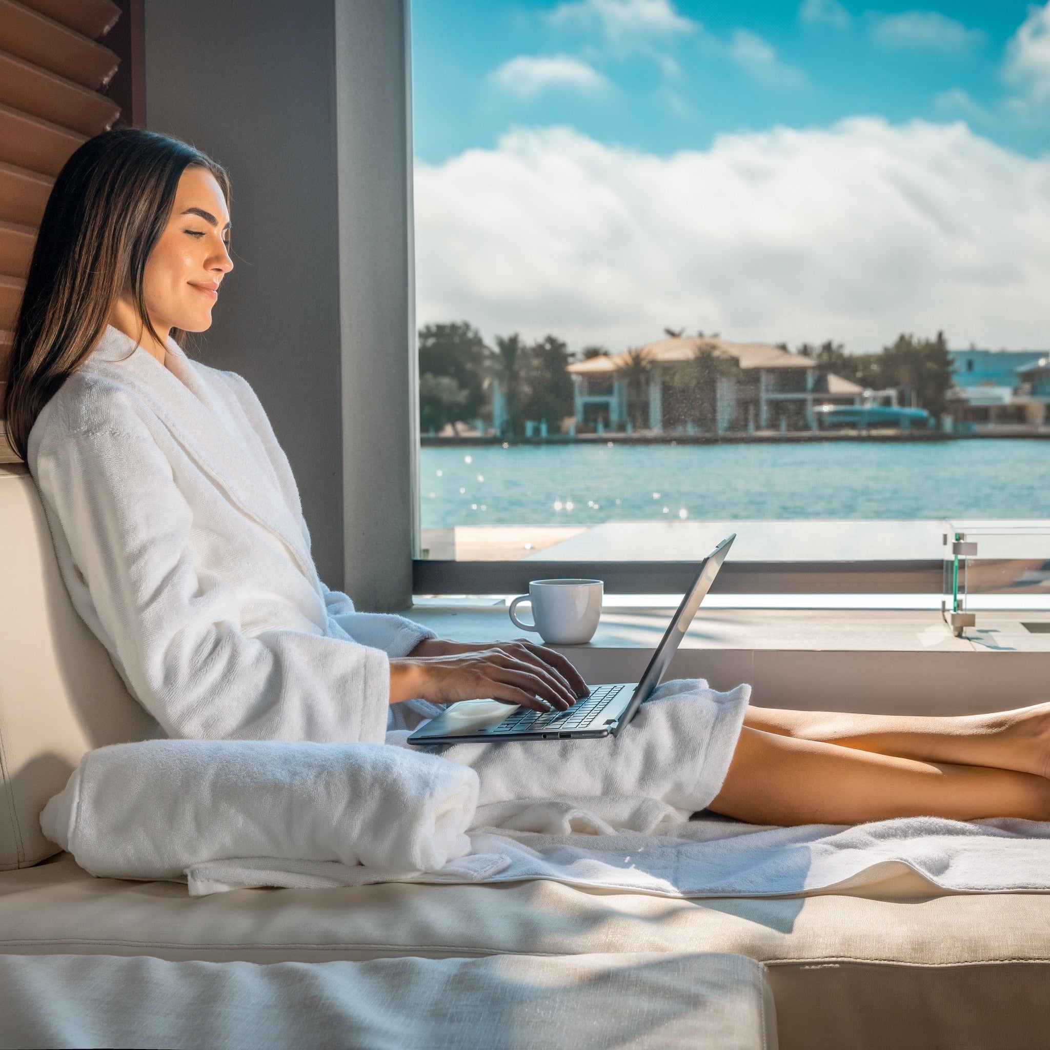 Woman in a white robe using a laptop by a large window with a scenic view.