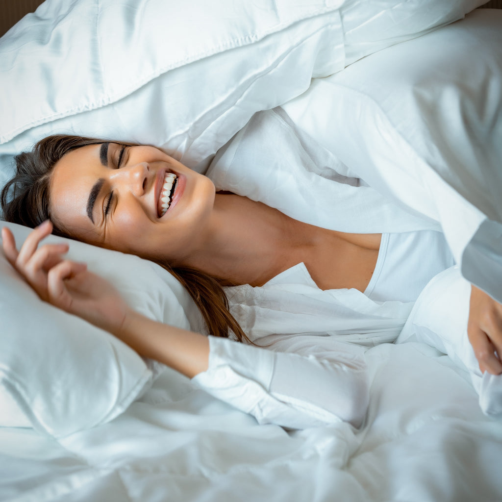 Woman laughing while laying in bed covering up with a white comforter.