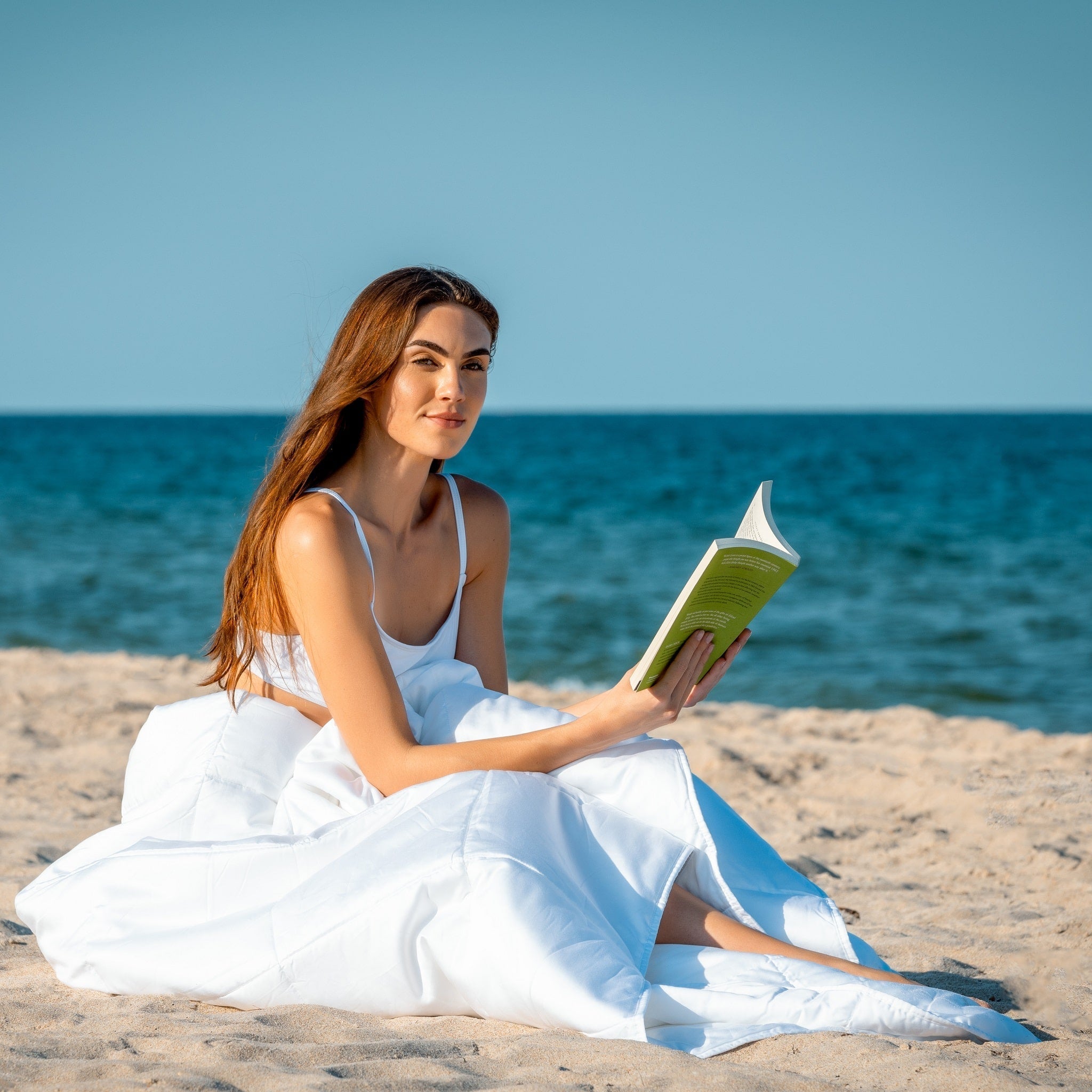 Woman sitting on beach reading a book wrapped up in a white comforter.