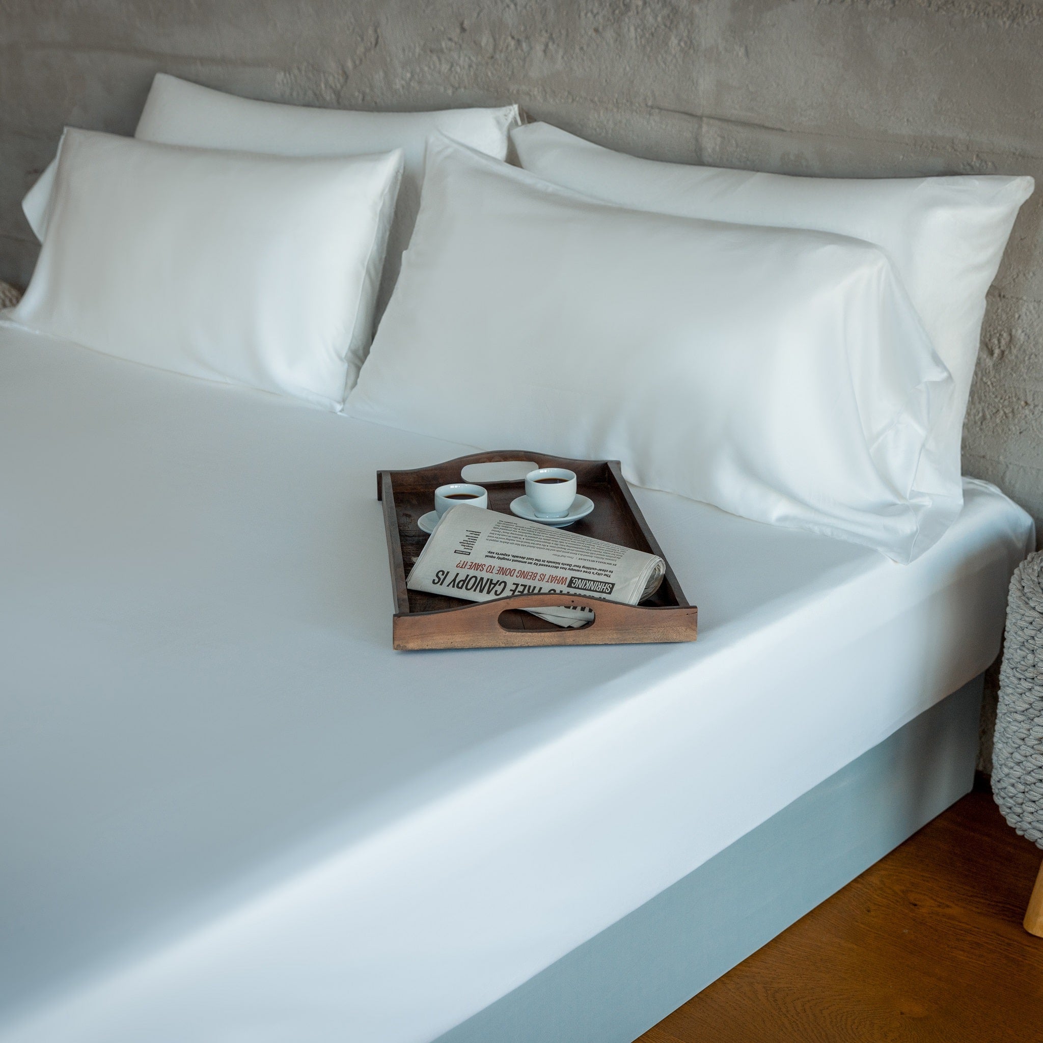 White pillows on a bed with a wooden tray holding cups and a newspaper.
